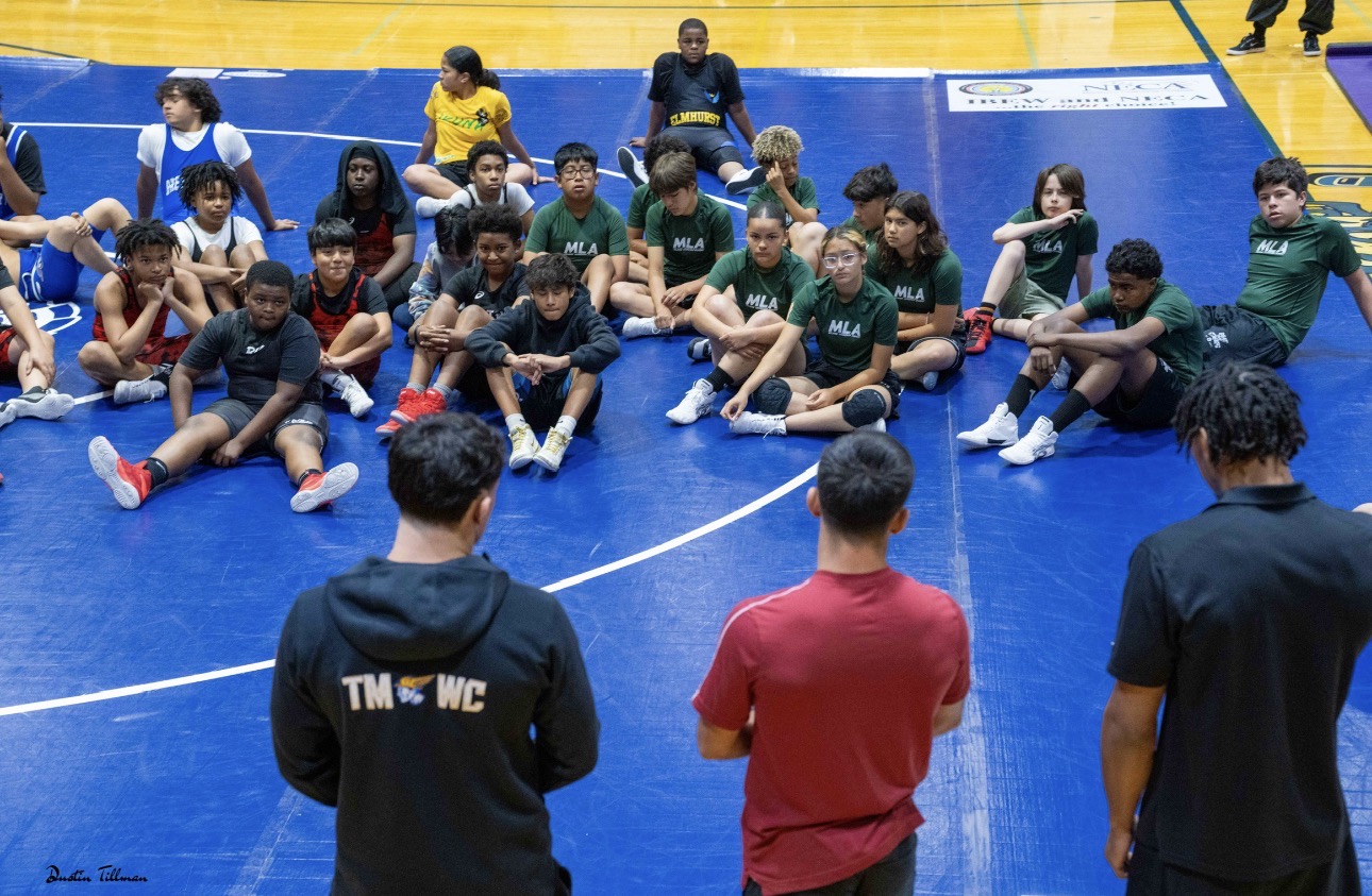 Group of young wrestlers sitting on the mat listening to coaches at a camp or clinic